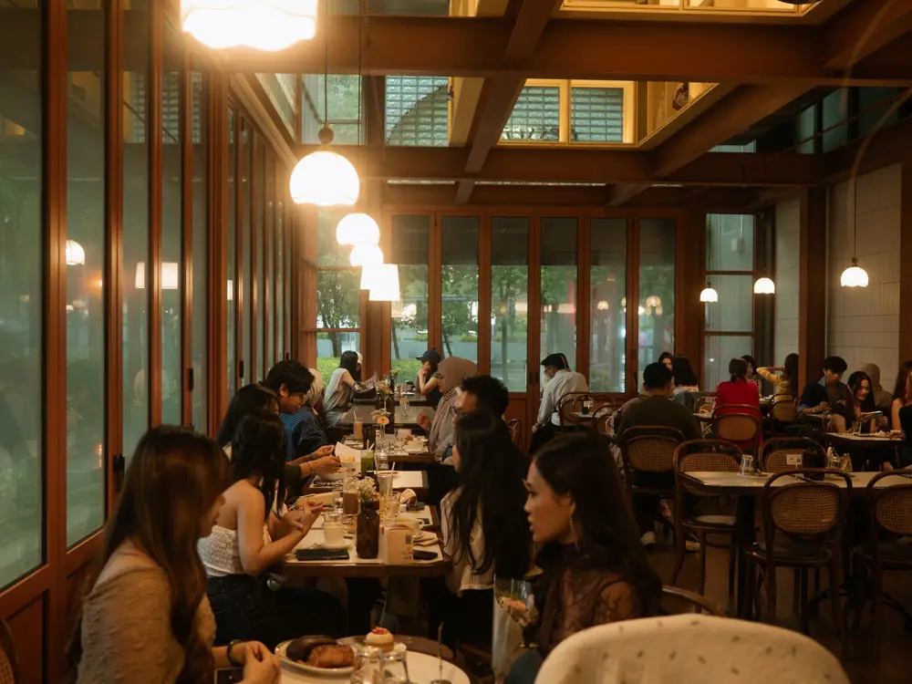 Diners at wooden tables in a warm, lamp-lit restaurant with tall windows